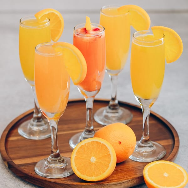 Five Mikayla's Cafe mimosas, some garnished with orange slices, on a wooden tray with fresh oranges.