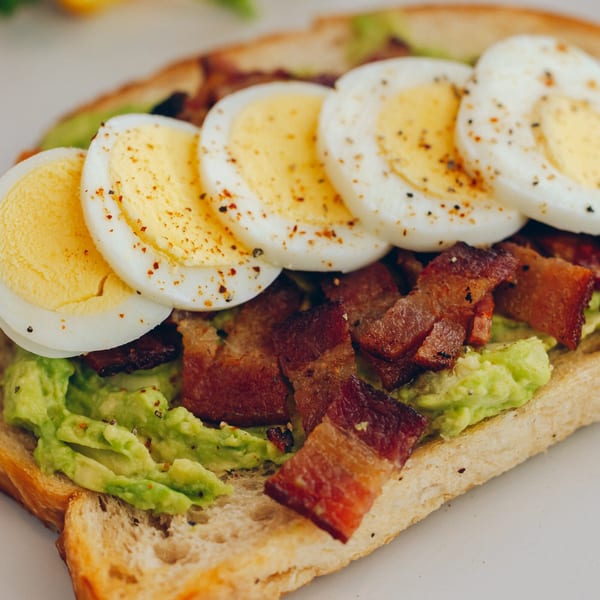 Mikayla's Cafe avocado toast with sliced hard-boiled eggs and crispy bacon, seasoned with pepper.