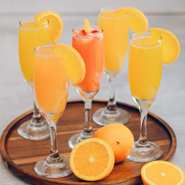 Five Mikayla's Cafe mimosas, some garnished with orange slices, on a wooden tray with fresh oranges.