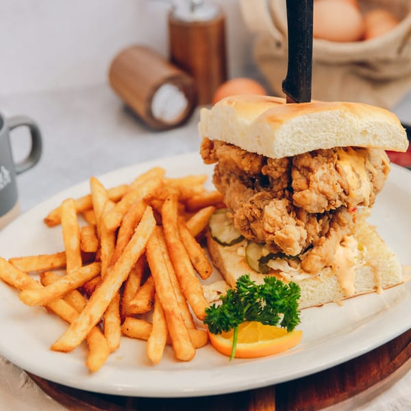 Mikayla's Cafe fried chicken sandwich with fries, a knife, and an orange slice on a white plate.