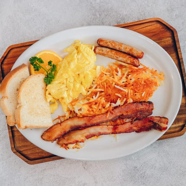 A Mikayla's Cafe breakfast plate with scrambled eggs, bacon, sausage, hash browns, toast, and orange slice.