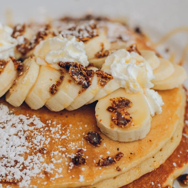 Fluffy pancakes topped with sliced bananas, whipped cream, granola, powdered sugar, and syrup.