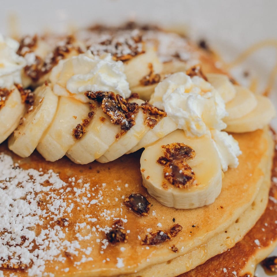 Fluffy pancakes topped with sliced bananas, whipped cream, granola, powdered sugar, and syrup.