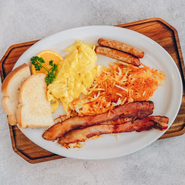 A Mikayla's Cafe breakfast plate with scrambled eggs, bacon, sausage, hash browns, toast, and orange slice.