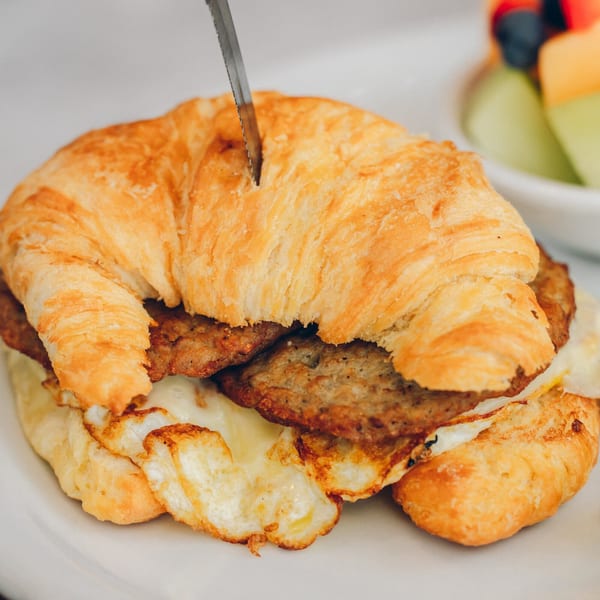 A knife cutting into a croissant breakfast sandwich with sausage and egg, with fruit in the background.