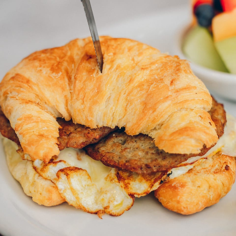 A knife cutting into a croissant breakfast sandwich with sausage and egg, with fruit in the background.
