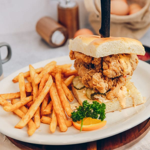 Crispy fried chicken sandwich with pickles, sauce, fries, parsley, and orange slice on a white plate.